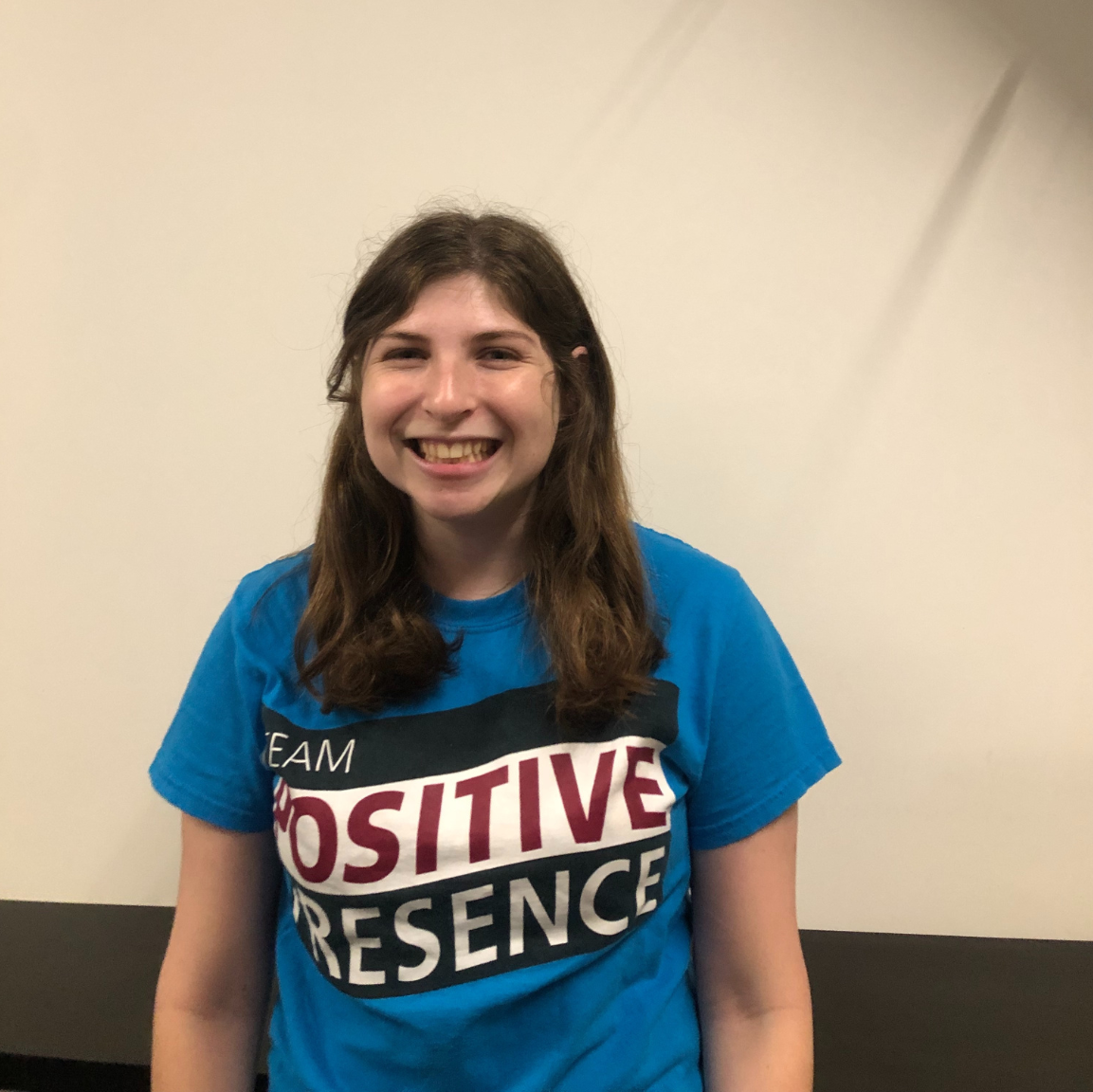 A white woman with brown hair standing in front of a blank wall, smiling widely in a tee shirt that reads "Team Positive Presence"..